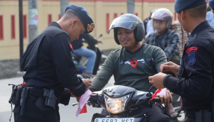 Personel Brimob Polda Banten membagikan dan memasang bendera Merah Putih kepada pengendara motor dalam rangka menyambut HUT ke-80 RI.